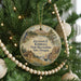 A round ornament hanging on a Christmas tree, featuring a scenic landscape with a cabin, mountains, and a bird.
