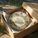 A round ornament in a wooden box, featuring a scenic landscape with a house, trees, and a boat, along with the text "JACKSON F. KIMBALL State Recreation Site, Oregon".