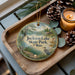A decorative ornament with a scenic lake and forest landscape, featuring the text "Jackson Lake State Park, Ohio". The ornament is placed on a wooden tray, with pine cones and a lit candle nearby.