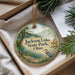 A decorative ornament with a scenic lake and park landscape, featuring a bird and a cabin, is displayed in a box with pine branches.