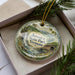 A decorative ornament in a box, featuring a scenic landscape with a waterfall and a wooden bridge, with the text "Jamaica State Park, Vermont" printed on it.