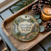 A decorative ornament featuring a scenic landscape with a waterfall, pine trees, and a castle, placed on a wooden tray alongside pine cones and a lit candle.