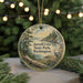 A circular ornament with a scenic landscape of a lake, forest, and mountains, featuring a bird in flight, is displayed in front of a blurred background with twinkling lights.