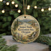 A round ornament with a scenic landscape of a mountain, river, and cabin, surrounded by pine trees and greenery, hangs in front of a blurred background of Christmas lights.
