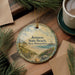 A decorative ornament featuring a beach scene with a lighthouse, surrounded by pine branches and a cup of coffee on a wooden surface.