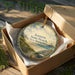 A round ornament in a box, depicting a scenic beach landscape with a lighthouse and a cloudy sky.