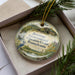 A decorative ornament in a box, featuring a blue jay and a scenic landscape with a building, surrounded by greenery.