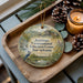 A decorative ornament with a bird perched on a branch, featuring the text "Jennings Environmental Education Center, Pennsylvania" on it, placed on a wooden tray alongside pine cones and a lit candle.