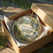 A decorative ornament in a box, featuring a scenic landscape with a waterfall and forest, with the text "Jenny Jump State Forest, New Jersey" printed on it.