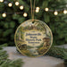 A circular ornament with a painting of a scenic landscape, featuring a river, buildings, and trees, hangs in front of a blurred background of Christmas lights.