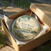 A round ornament in a box, featuring a lighthouse and beach scene, with the text "Jones Beach State Park, New York" printed on it.