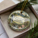 A decorative ornament in a box, featuring a scenic landscape with a waterfall, pine trees, and a bridge.