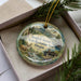 A decorative ornament with a scenic landscape featuring a river, mountains, and wildlife, including a deer, is displayed in a box with pine branches.