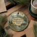 A round ornament with a scenic landscape featuring a deer, mountains, and a cabin, hanging on a wooden surface.