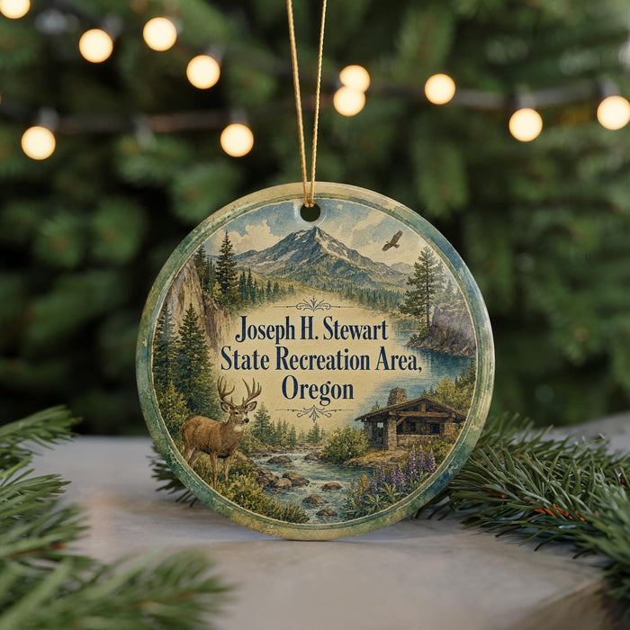A round ornament featuring a scenic landscape with a mountain, river, and cabin, along with a deer and pine trees, is displayed on a table with a blurred background of Christmas lights.