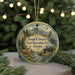 A round ornament featuring a scenic landscape with a mountain, river, and cabin, along with a deer and pine trees, is displayed on a table with a blurred background of Christmas lights.
