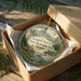 A round ornament in a wooden box, featuring a scenic landscape with a deer, mountains, and a stream.