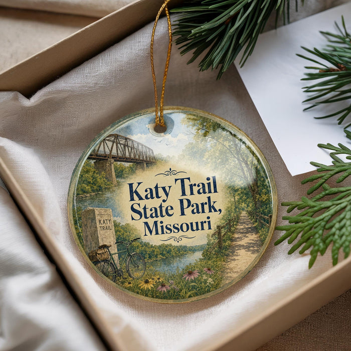 A decorative ornament with a scenic image of a trail and bridge at Katy Trail State Park, Missouri, placed in a box with pine branches.