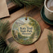A round ornament with a scenic image of a trail in Missouri, featuring a bridge and pine trees, is displayed on a wooden surface alongside a cup of coffee and some papers.