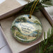 A decorative ornament in a box, featuring a scenic landscape with a house, trees, and a body of water.