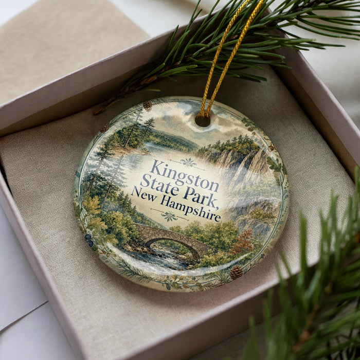 A decorative ornament in a box, featuring a scenic landscape with a bridge and pine trees, with the text "Kingston State Park, New Hampshire" printed on it.