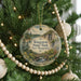 A round ornament hanging on a Christmas tree, featuring a scenic landscape with a bridge and pine trees.