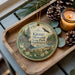 A decorative ornament with a scenic image of Kitsap Memorial State Park, Washington, sits on a wooden tray alongside pine cones and a lit candle.