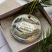 A decorative ornament in a box, featuring a scenic landscape with mountains, waterfalls, and pine trees.