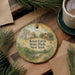 A decorative ornament with a rural scene and text, placed on a wooden surface next to a cup of coffee and some pine branches.