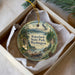 A decorative ornament in a box, featuring a landscape scene with mountains, pine trees, and a bird in flight.
