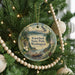 A round ornament hanging on a Christmas tree, featuring a scenic landscape with mountains, pine trees, and a bird in flight.