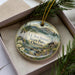 A decorative ornament in a box, featuring a scenic landscape with mountains, waterfalls, and a small cabin.