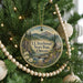 A round ornament hanging on a Christmas tree, featuring a scenic landscape with mountains, a river, and a cabin.