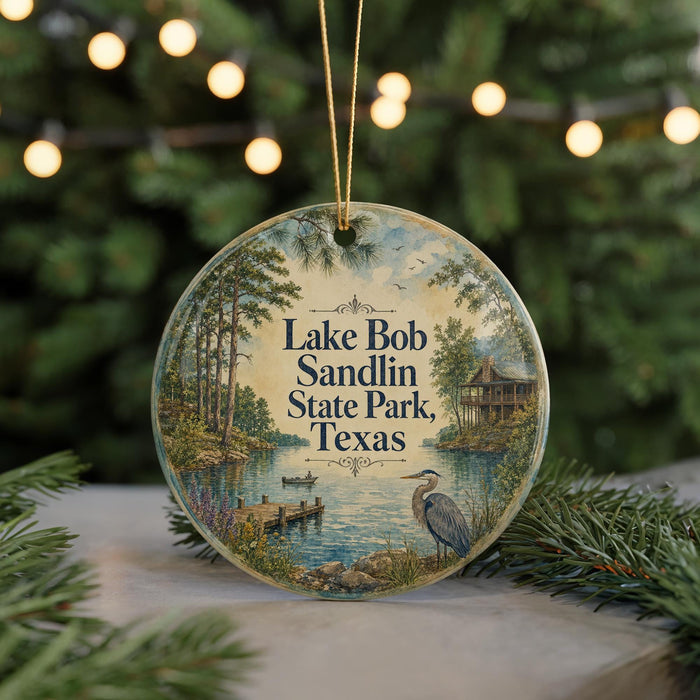 A round ornament with a painting of Lake Bob Sandlin State Park in Texas, featuring a bird and a dock, is displayed on a table with pine branches in the background.