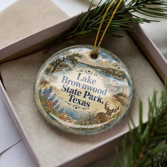 A decorative ornament in a box, featuring a scenic image of Lake Brownwood, Texas, with a deer in the foreground.