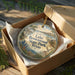 A circular ornament in a wooden box, featuring a scenic landscape with a lake, trees, and a deer.