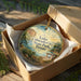 A decorative ornament in a box, featuring a scenic landscape with a lake, mountains, and a bridge.