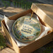 A decorative ornament in a wooden box, featuring a scenic landscape with mountains, trees, and a lake.