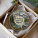 A decorative ornament in a box, featuring a scenic landscape with mountains, pine trees, and a lake.