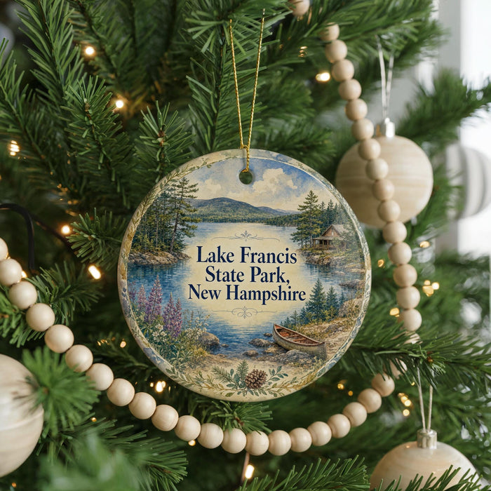 A round ornament hanging on a Christmas tree, featuring a scenic landscape with a lake, mountains, and a cabin.