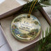 A decorative ornament in a box, featuring a scenic landscape with a lake, trees, and a cabin.