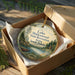 A round ornament with a scenic lake and forest landscape, placed in a cardboard box with a gold string.