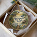 A decorative ornament in a box, featuring a scenic landscape with a lake, trees, and a cabin, along with a bird and a deer.