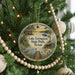 A round ornament hanging on a Christmas tree, featuring a scenic landscape with a lake, trees, and a cabin.