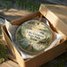 A decorative ornament in a box, featuring a scenic landscape with a lake, trees, and wildlife, including a bird and a deer.