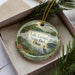 A decorative ornament with a lake and forest scene, featuring a bird and a boat, is displayed in a box with pine branches.
