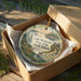 A decorative ornament in a box, featuring a scenic landscape with a lake, trees, and a deer.