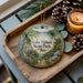 A wooden tray with a decorative ornament featuring a scenic landscape of Lake Milton State Park, Ohio, along with pine cones and a lit candle.