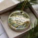 A decorative ornament in a box, featuring a scenic landscape with a lake, minerals, and a bird.