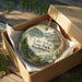A round ornament in a wooden box, featuring a scenic landscape with a lake, mountains, and trees.